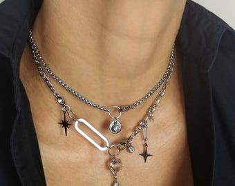 Könnte beinhalten: Eine mehrschichtige silberne Halskette mit verschiedenen Anhängern. Die Halskette hat eine Kette, einen Stern, einen Tropfenanhänger und einen kreisförmigen Anhänger mit Monddesign. Die Halskette wird über einem schwarzen Hemd getragen.