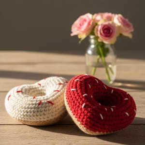 May include: Two heart-shaped crochet donuts, one cream-colored with red stitching and the other red with white stitching. The donuts sit on a wooden surface, with a vase of pink roses in the background.