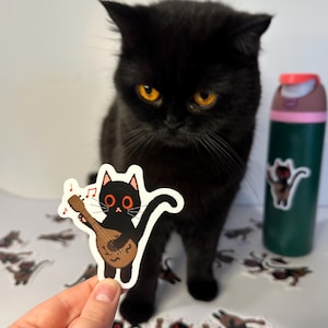 May include: A black cat with orange eyes looks at a sticker of a black cat playing a lute. The sticker has red eyes and is on a white background. The cat is holding a brown lute and there are musical notes above its head.