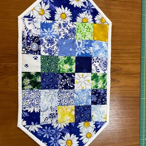 May include: A quilted table runner with a patchwork design. The runner features a variety of blue and white floral patterns, including daisies, and is bordered with a white trim. The runner is approximately 20 inches long.