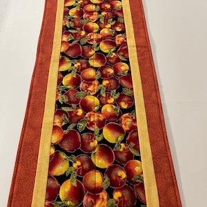 May include: A rectangular table runner featuring a pattern of red and yellow apples with green leaves on a black background. The runner has a red border with a floral design and a yellow stripe.