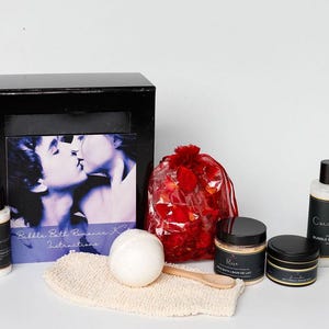 Puede incluir: Un set de caja negra con temática romántica, que incluye un libro, productos de baño y una bolsa de pétalos de rosa rojos. El set incluye una bomba de baño, una cuchara de madera y un paño. Los productos están en recipientes negros.