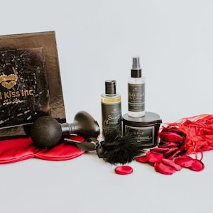 以下が含まれることがあります： 黒い箱、アイマスク、さまざまな製品ボトルが入ったギフトセット。箱には「Red Kiss Inc」の文字とハートのロゴが入っています。その他のアイテムには、黒い羽、黒い球根、赤いバラの花びらなどがあります。