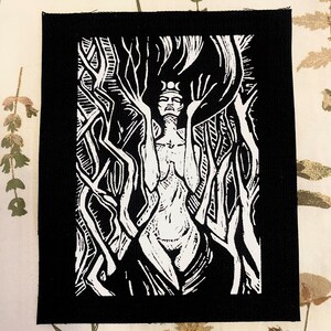 May include: Black and white embroidered patch featuring a stylized image of a woman with a crown and a crescent moon on her head, standing within a forest of trees.