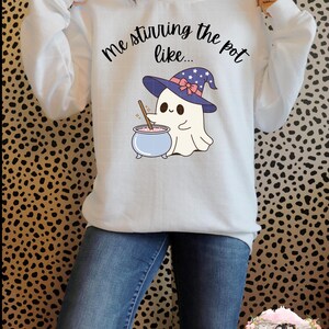 May include: White sweatshirt with a ghost wearing a witch hat and stirring a cauldron. The text on the shirt reads "Me stirring the pot like..."