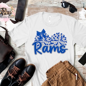 Può includere: Maglietta bianca con un design a pompon blu e grigio e la parola "Rams" in lettere blu.