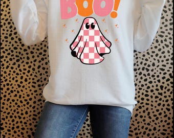 May include: White long-sleeve sweatshirt featuring a Halloween-themed design. The design includes the word "Boo!" in pink and orange, along with a checkered pink and white ghost graphic. Small orange stars are scattered around the ghost.