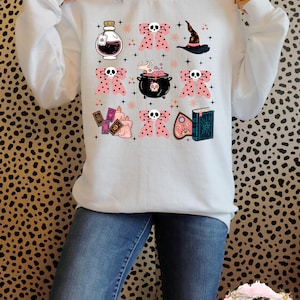 Pode incluir: Camisola branca com um design rosa e preto com um chapéu de bruxa, caldeirão, frascos de poções, cartas de tarô e outros elementos de bruxaria.