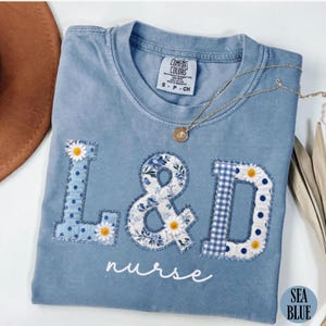 May include: Sea blue Comfort Colors t-shirt with the text "L & D nurse". The letters are appliqued with blue floral, polka dot, and gingham patterns, and daisy accents. A gold necklace with a pendant rests on the shirt.