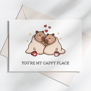 May include: A greeting card with an illustration of two capybaras under a blanket, with hearts above. The card reads "YOU'RE MY CAPPY PLACE". Two mugs are also shown. The card is on a white background with a shadow.