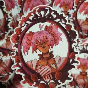 Op de afbeelding: Een sticker met een gestileerde illustratie van een personage in een decoratieve, sierlijke lijst. Het personage heeft roze haar en wordt omlijst door rode rozen. De lijst is donkerrood.