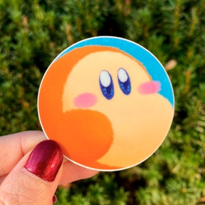 May include: A round sticker with a cartoon character with orange, yellow, and pink skin tones. The character has large blue eyes and a pink blush on its cheeks.