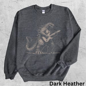 May include: Dark heather grey sweatshirt with a tan illustration of Godzilla playing an electric guitar. The text "Dark Heather" is at the bottom. The sweatshirt has a crew neck and long sleeves.
