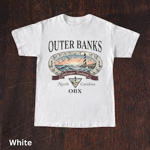 Puede incluir: Camiseta blanca con un dise&ntilde;o gr&aacute;fico colorido. El dise&ntilde;o incluye las palabras "Outer Banks" y "Saltwater Tradition", un faro, olas y p&aacute;jaros. Tambi&eacute;n est&aacute; presente el texto "North Carolina OBX".
