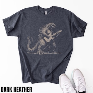 May include: Dark heather gray t-shirt featuring a Godzilla graphic playing a guitar. The illustration is in a neutral color, and the shirt is paired with white sneakers. The text "DARK HEATHER" is at the bottom.