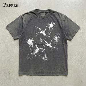 May include: A charcoal gray short-sleeve t-shirt featuring a white graphic print of three flying ravens in the center. The word Pepper is printed in the upper left corner in a serif font. The shirt has a crew neckline and a washed-out fabric finish.