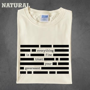 Könnte beinhalten: Cremefarbenes T-Shirt mit dem Text "everything is fine trust your government" in schwarzer, blockartiger Schrift. Das Wort "NATURAL" ist in der oberen linken Ecke des Shirts aufgedruckt.