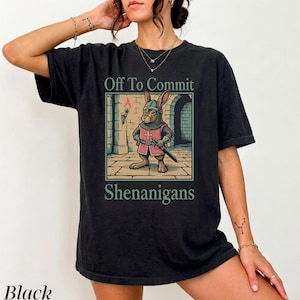 May include: A black short-sleeve t-shirt featuring a graphic of a rabbit dressed as a medieval knight in armour, holding a sword. The text above reads Off To Commit and below reads Shenanigans. The design includes a stone wall and a torch.