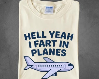 May include: Cream-colored t-shirt with the text "HELL YEAH I FART IN PLANES" in dark blue, along with a light purple airplane graphic. The shirt has a crew neck and a tag from GILDAN HAMMER.