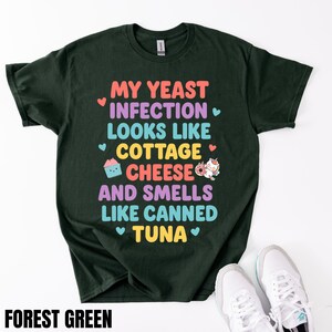 Puede incluir: Camiseta verde bosque con el texto "MY YEAST INFECTION LOOKS LIKE COTTAGE CHEESE AND SMELLS LIKE CANNED TUNA" en letras coloridas. La camiseta tiene cuello redondo y mangas cortas. Zapatillas blancas en la parte inferior de la imagen.