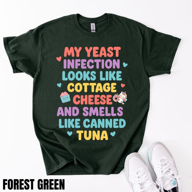 Puede incluir: Camiseta verde bosque con el texto "MY YEAST INFECTION LOOKS LIKE COTTAGE CHEESE AND SMELLS LIKE CANNED TUNA" en letras coloridas. La camiseta tiene cuello redondo y mangas cortas. Zapatillas blancas en la parte inferior de la imagen.