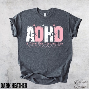 Könnte beinhalten: Ein dunkelgrau meliertes Kurzarm-T-Shirt mit dem Text ADHD in einer abgenutzten weißen und rosa Schriftart und drei kleinen Sternsymbolen darüber. Unter dem Text steht der Satz A Diva Has Distractins in Rosa mit einer gewellten Linie.