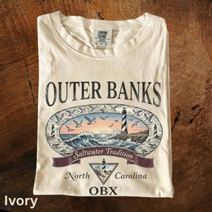 Puede incluir: Camiseta color marfil con un gr&aacute;fico de Outer Banks, con un faro, olas y gaviotas. El texto incluye "Outer Banks", "Saltwater Tradition", "North Carolina" y "OBX".