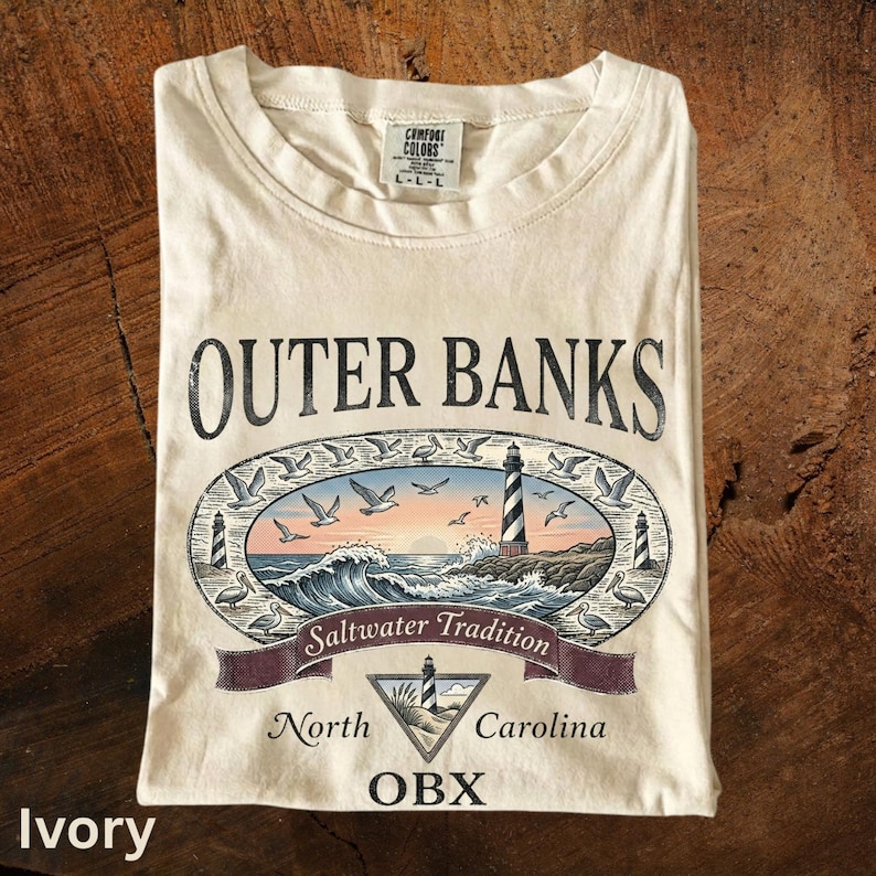 Puede incluir: Camiseta color marfil con un gr&aacute;fico de Outer Banks, con un faro, olas y gaviotas. El texto incluye "Outer Banks", "Saltwater Tradition", "North Carolina" y "OBX".