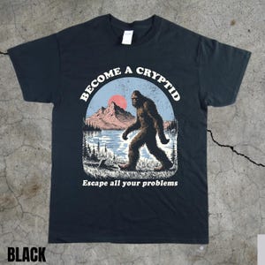 Puede incluir: Camiseta negra con un gráfico de un Bigfoot caminando cerca de un lago y montañas, con el texto "BECOME A CRYPTID" y "Escape all your problems". El diseño presenta un sol rojo y un cielo azul.