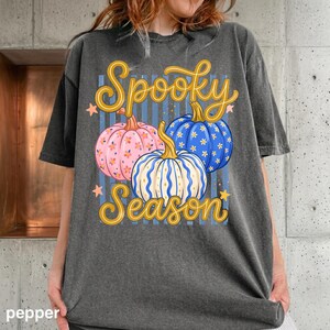 May include: A charcoal gray t-shirt featuring a graphic of three pumpkins in pink, blue, and white with floral patterns. The text Spooky Season is printed in yellow cursive script above and below the pumpkins, accented by small orange stars.