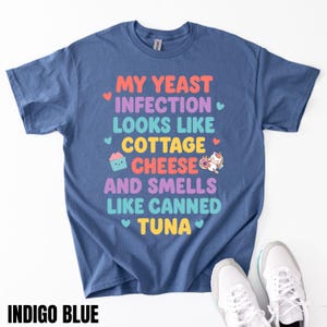 Puede incluir: Camiseta azul &iacute;ndigo con el texto "MY YEAST INFECTION LOOKS LIKE COTTAGE CHEESE AND SMELLS LIKE CANNED TUNA" en letras coloridas. La camiseta presenta una magdalena y un personaje de dibujos animados con un donut.