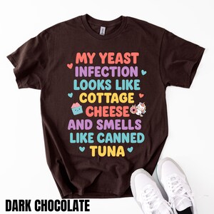 Puede incluir: Camiseta marr&oacute;n chocolate oscuro con el texto "MY YEAST INFECTION LOOKS LIKE COTTAGE CHEESE AND SMELLS LIKE CANNED TUNA" en letras coloridas. Tambi&eacute;n se muestra una magdalena y un donut de unicornio.