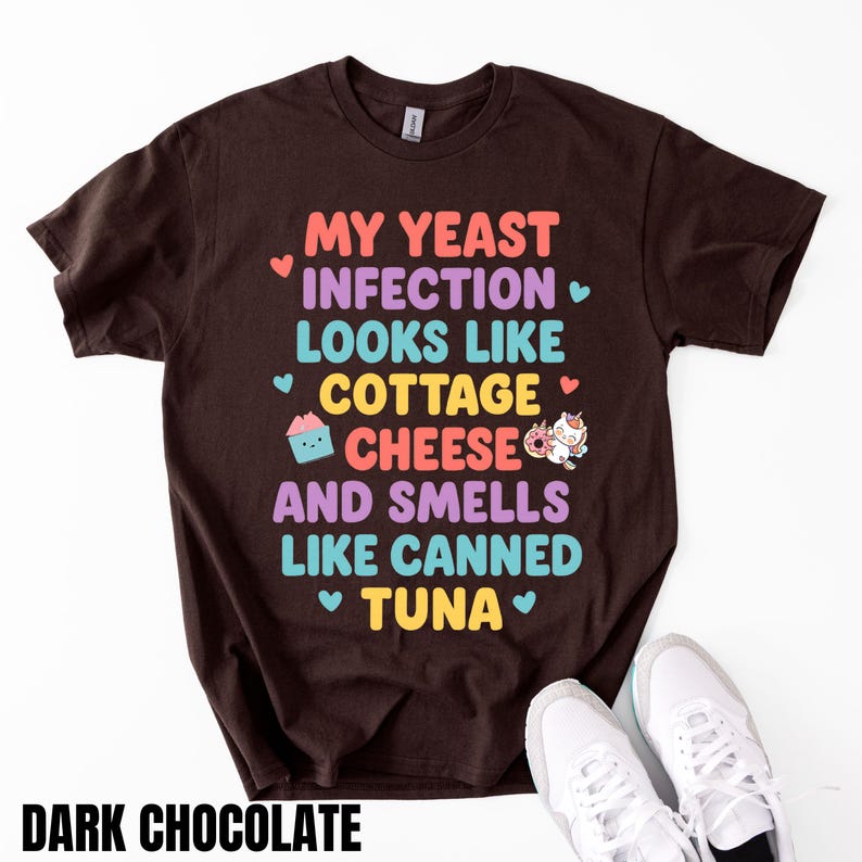 Puede incluir: Camiseta marr&oacute;n chocolate oscuro con el texto "MY YEAST INFECTION LOOKS LIKE COTTAGE CHEESE AND SMELLS LIKE CANNED TUNA" en letras coloridas. Tambi&eacute;n se muestra una magdalena y un donut de unicornio.