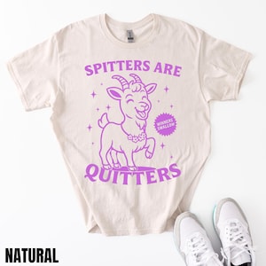 May include: A natural-colored t-shirt with a purple graphic of a smiling goat and the text "SPITTERS ARE QUITTERS". The shirt also features the text "WINNERS SWALLOW". White sneakers are at the bottom.
