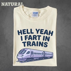 May include: A natural-coloured t-shirt featuring the dark blue text "HELL YEAH I FART IN TRAINS" printed above a graphic illustration of a light purple passenger train travelling on tracks. The shirt has a crew neckline and a Gildan brand label.
