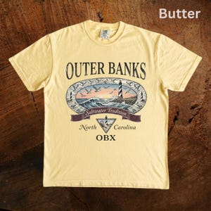 Puede incluir: Camiseta amarilla mantequilla con un dise&ntilde;o de Outer Banks. El gr&aacute;fico incluye un faro, olas y el texto "Saltwater Tradition, North Carolina, OBX". La camiseta est&aacute; hecha de un material suave.