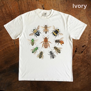 Könnte beinhalten: Elfenbeinfarbenes T-Shirt mit einem kreisförmigen Design von Bienenillustrationen in Braun, Schwarz, Gelb und Grün. Das Shirt ist elfenbeinfarben, und das Wort "Ivory" ist oben rechts aufgedruckt.