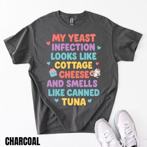 Puede incluir: Camiseta de color carb&oacute;n con el texto "MY YEAST INFECTION LOOKS LIKE COTTAGE CHEESE AND SMELLS LIKE CANNED TUNA". El texto est&aacute; en varios colores y fuentes. Una magdalena y un unicornio tambi&eacute;n aparecen en la camiseta.