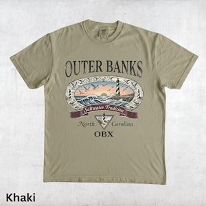 Puede incluir: Camiseta caqui con un dise&ntilde;o gr&aacute;fico de Outer Banks, Carolina del Norte. El dise&ntilde;o incluye un faro, olas, gaviotas y el texto "Saltwater Tradition" y "OBX".