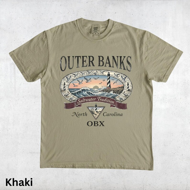 Puede incluir: Camiseta caqui con un dise&ntilde;o gr&aacute;fico de Outer Banks, Carolina del Norte. El dise&ntilde;o incluye un faro, olas, gaviotas y el texto "Saltwater Tradition" y "OBX".
