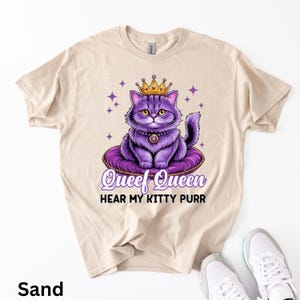 May include: Sand-colored t-shirt featuring a purple cat wearing a gold crown and necklace, sitting on a purple cushion. The text "Queef Queen Hear My Kitty Purr" is below the cat. The shirt is paired with white sneakers.