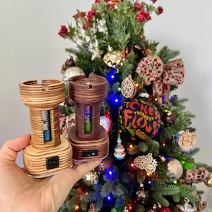 Könnte beinhalten: Zwei Holz-Sanduhren-Ornamente mit einem Lichtschalter an der Unterseite. Die Sanduhren sind mit blauem und grünem Sand gefüllt. Die Ornamente befinden sich vor einem geschmückten Weihnachtsbaum.