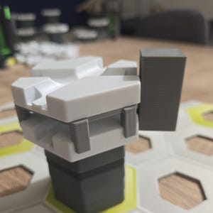 Op de afbeelding: Een wit en grijs 3D-geprint model van een gebouw met een grijze rechthoekige uitbreiding aan de zijkant. Het model staat op een witte en grijze zeshoekige tegel.