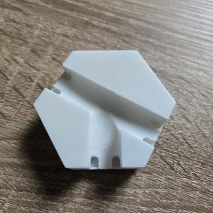 Könnte beinhalten: Weißes, sechseckiges Puzzle aus Kunststoff mit einem geschwungenen Kanal, der durch ihn verläuft. Das Puzzle hat drei Löcher auf einer Seite.