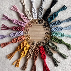 May include: A collection of 20 colourful macrame keychains with tassels. Each keychain features a unique pattern of knots and is attached to a silver key ring.