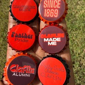 May include: Six circular bottle caps with serrated edges, featuring red and dark backgrounds with printed text including Orangeburg Raised Me, Since 1869, Panther Pride, Claflin Made Me, Claflin Alumni, and Pretty Girls Choose Claflin in various fonts.