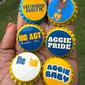 May include: Six circular bottle caps featuring blue and gold designs. Text includes GREENSBORO RAISED ME, NC A&T MADE ME, AGGIE PRIDE, BLUE & GOLD FOREVER, and AGGIE BABY. One cap features a cartoon character wearing a blue hoodie and yellow pants.