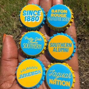 May include: Six circular metal bottle caps featuring blue and yellow color schemes with printed text. The caps display phrases including SINCE 1880, BATON ROUGE MADE ME, Pretty Girls Choose SOUTHERN, SOUTHERN ALUMNI, HUMAN JUKEBOX, and Jaguar NATION.