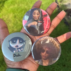May include: Three circular pin-back buttons featuring different printed images. One button shows a person holding a mug, another displays a cartoon character with the text "Duh.", and the third depicts a person wearing sunglasses and a hat.