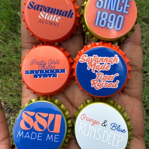 May include: Six circular metal bottle caps featuring various designs related to Savannah State University. The caps display text including Savannah State Alumni, Since 1890, Pretty Girls Choose Savannah State, Savannah Made Tiger Raised, SSU Made Me, and Orange & Blue Runs Deep in orange, blue, and white colors.